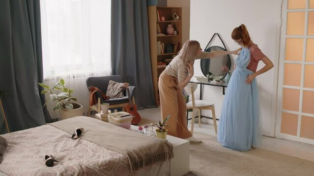 Mature mother standing in front of her teenage daughter and helping her try on long blue fatin evening dress while both preparing for prom celebration before graduation event