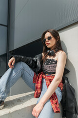 Stylish beautiful young woman with cool sunglasses with headphones in fashion urban clothes with a leather jacket, bandana top and jeans sitting in the city near a building