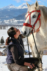 A woman in sunglasses looks at a white horse against the backdrop of mountains in winter