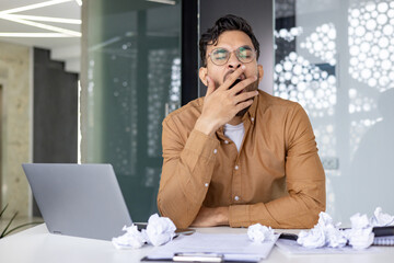 Tired businessman yawning at desk with laptop and crumpled papers in modern office