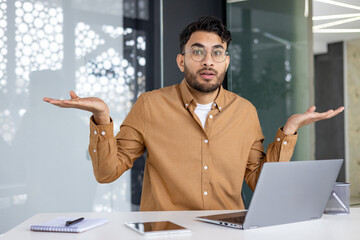 Confused young man in office with laptop, unsure expression and hand gesture