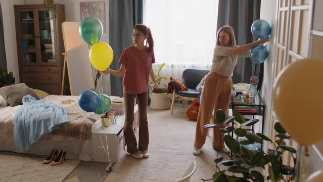 Teenage girl putting bunch of balloons on the floor by bed and looking at her mother pinning decorations to wall while both preparing domestic room for celebrating prom