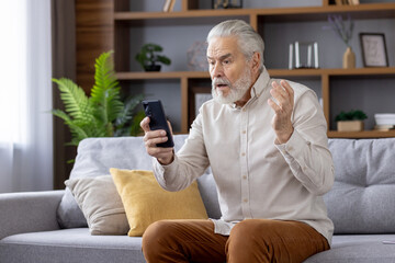 Shocked senior man sitting on sofa at home and looking surprised at phone screen, got bad news, spreads hands