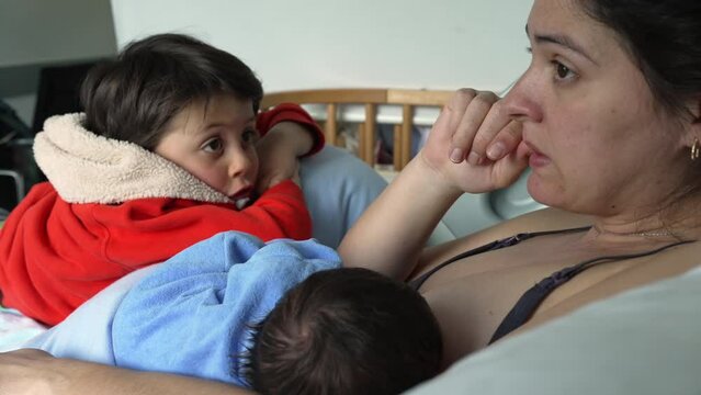 Mother breastfeeding newborn while older child leans on her, conveying the warmth and closeness of family life and the support that older siblings provide during early parenting