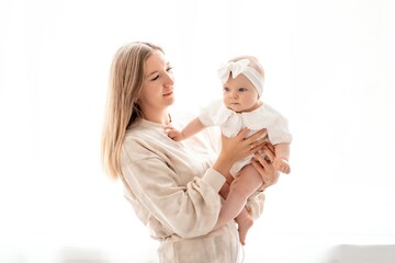 a mother with a newborn baby in her arms at home on a white background of a bed or window, a space for text, a young mother gently kisses and hugs her baby, maternal love