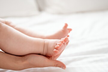 Mom's hand holds the small legs of her newborn baby on the white background of the bed, space for...