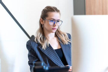 A beautiful girl is sitting at a computer monitor wearing glasses and a classic striped suit. A businesswoman is focused on solving problems. Selected focus.