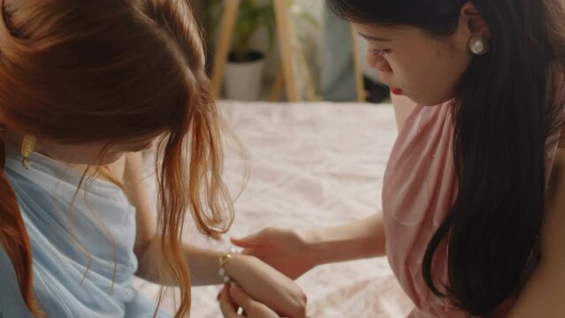 Tilt up of two teenage girls wearing pink and blue evening dresses sitting on bed at home and trying on accessories while one of them putting pearl bracelet on wrist of her friend