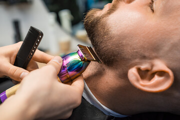 Barber cuts man beard creating stylish shape with trimmer in barbershop closeup. Hairdresser works...