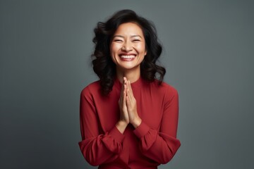 Fototapeta premium Portrait of a smiling asian woman in her 40s joining palms in a gesture of gratitude while standing against blank studio backdrop