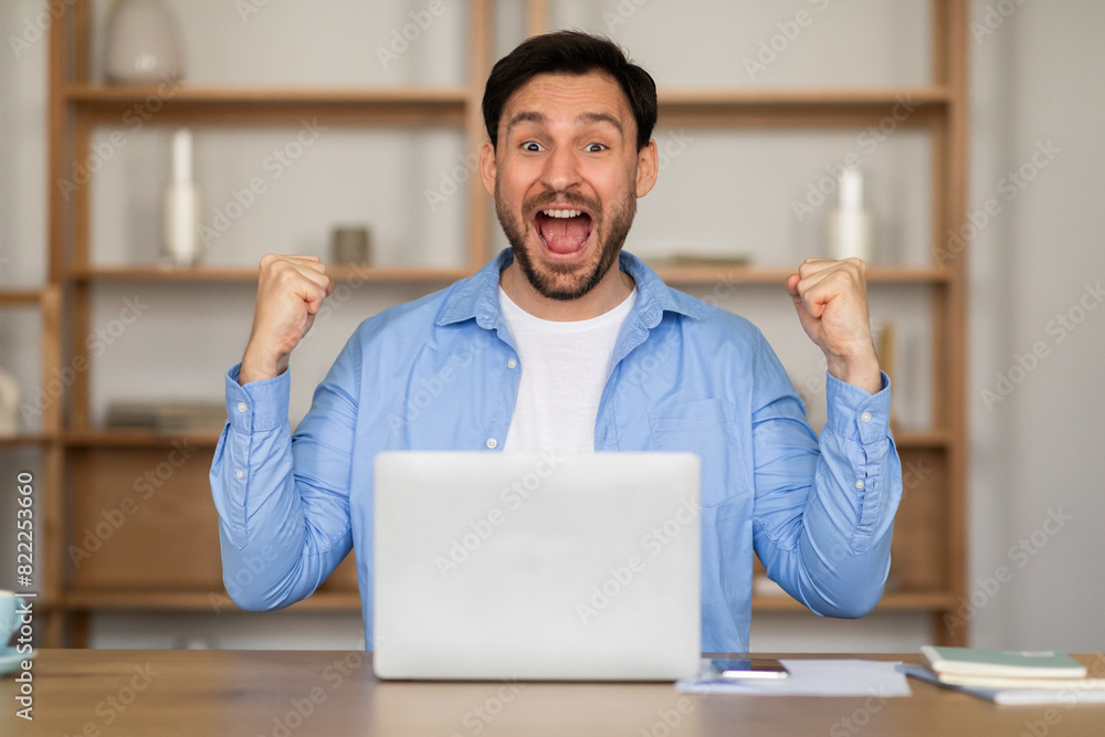 Poster Excited man is seated at a table with a laptop open in front of him. He appears focused on the screen, possibly working or studying, clenching fists and exclaiming