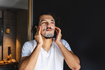 latin man applying exfoliating facial mask treatment and taking care of skin in loft apartment in Mexico Latin America, hispanic people at home	