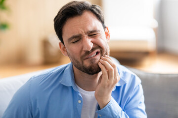 A man is shown sitting on a couch, visibly in discomfort, holding his jaw with a pained expression...