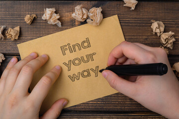 A hand is writing on a piece of paper that says find your way. The paper is on