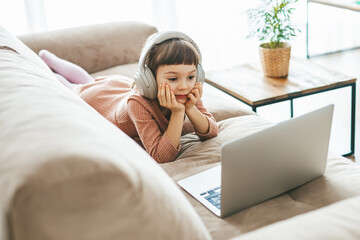 A cute little girl, aged 5-6 years old, wearing headphones while captivated by the laptop screen....