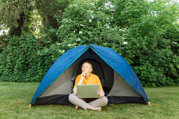 Woman working on laptop near camping tent outdoors surrounded by beautiful nature