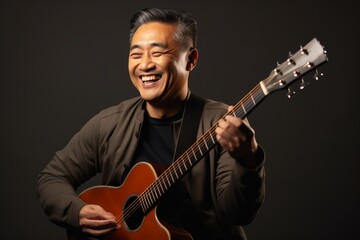 Portrait of a satisfied asian man in his 50s playing the guitar while standing against blank studio backdrop
