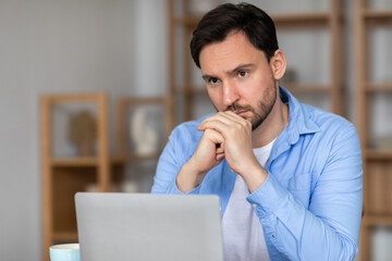 A man is seated in front of a laptop, deep in thought, with his hand resting on his chin. He...