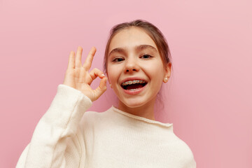 cheerful teenage girl with braces shows ok gesture on pink isolated background, child approves and...