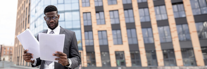Young black man in suit checking reports or getting ready for job interview, holding documents or CV, standing outdoors against office center, copy space