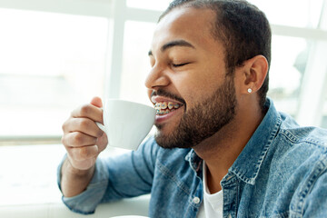 African American man with braces drinks coffee from a cup and smiles in a white cafe, a man with stubble drinks tea from a white mug