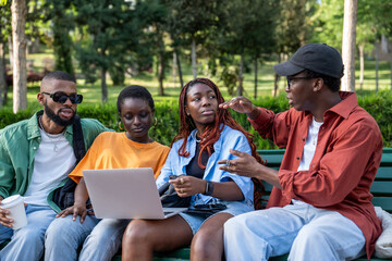Focused african american student talking with friends sitting with laptop on bench in college campus. Group of interested black girls and guys sharing ideas for study project brainstorming, discussing