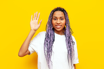 cheerful african american woman with colored dreadlocks greets and waves on yellow isolated...