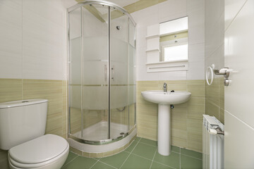 A bathroom with tiles in various shades of green and white, white wooden doors, a shower cabin with glass screens and white porcelain toilets