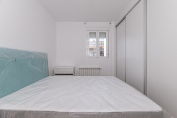 Bedroom with uninstalled bed with protective plastics on, built-in wardrobe and aluminum window in the background