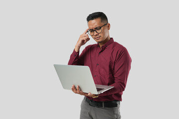 A young handsome Asian man in a suit wearing glasses and making thinking expressions and holding a laptop.