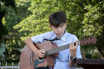 Asian preteen schoolboy in school uniform sitting and playing acoustic guitar in schoolpark,...