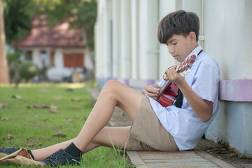 Asian preteen schoolboy in school uniform sitting near cream wall and playing ukulele after school.