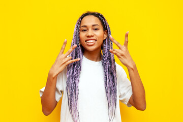 cheerful african american woman with colored dreadlocks shows rap gesture with fingers on yellow...