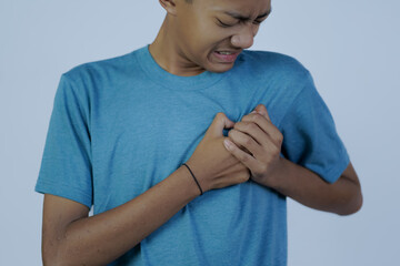 close up portrait of young Asian man in blue shirt moaning in pain while holding his chest, heart disease, isolated in gray background.