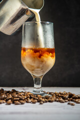 Rice milk being poured over french press coffee in a clear glass, standing on a heap of roasted coffe beans, a white wood surface with a black textured background.