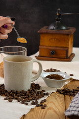 Hand holding a spoon with brown sugar in a cup of coffe sitting in the medium ground on a heap of roasted coffee beans, with a  jar of brown sugar and a small bowl with grinded coffee. In the backgrou