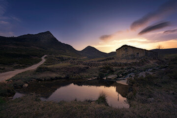 warm sunrise at the Pardo waterfall, in the mountain pass of Estacas de Trueba, Burgos, with a small pond in the foreground and a pasiega cabin between mountains