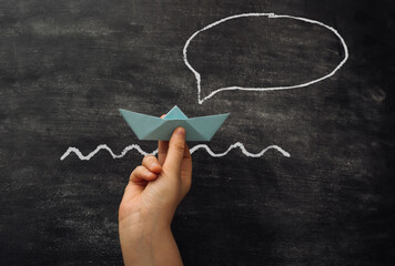 A hand holding a paper boat in front of a chalkboard with a speech bubble