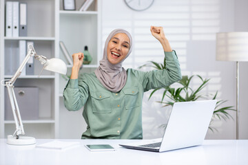 A cheerful businesswoman wearing a hijab celebrating success at her desk in a modern office. She...