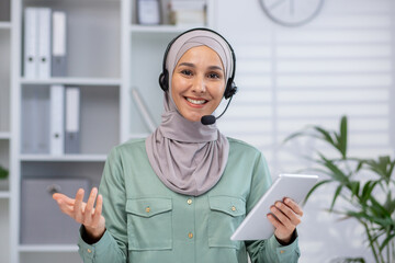 Confident woman wearing hijab and headset working in a modern office setting. She holds a tablet...
