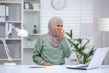 Businesswoman yawning in front of laptop, showing fatigue in a bright and modern office environment.
