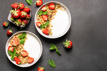 Fresh Greek Yogurt with granola and strawberries decorated with aromatic mint leaves on black stone background top view, copy space for your design. Healthy breakfast concept.