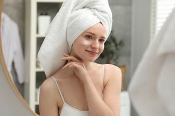 Beautiful woman with freckles and cream on her face near mirror in bathroom