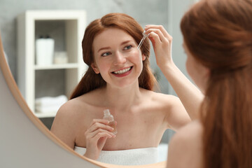 Smiling woman with freckles applying cosmetic serum onto her face near mirror in bathroom