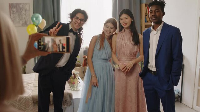 Hand of mature woman with smartphone standing in front of group of intercultural teenage graduates of highschool and taking picture or recording video of them at prom party