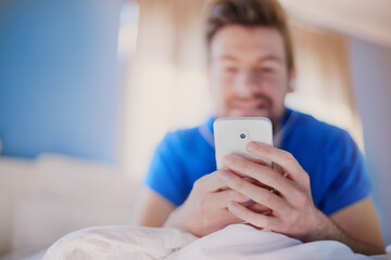 Hands, phone and man in bedroom, morning and smile for communication, reading and mobile app in home. Person, smartphone and relax on bed with typing, scroll and check notification on social network - Powered by Adobe