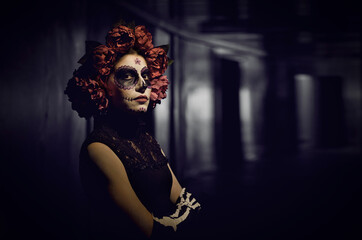 Portrait of woman dressed for Halloween or Day of the Dead. Lady with rose wreath and Catrina skull makeup standing on dark background inside dark building looking at camera with unfriendly expression