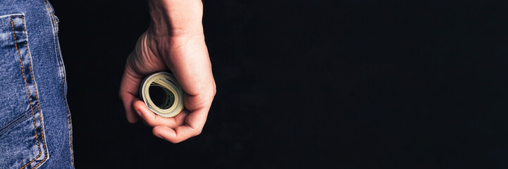 Male hand holding rolled-up money on dark background banner. Panoramic web header. Wide screen...