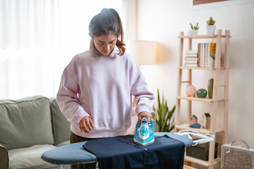 washing, cleaning, laundry, garment, steam, housewife, living, housekeeping, housework, hygiene. A woman is ironing a shirt in a living room. The room is furnished with a couch and a potted plant.