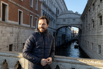 A man in casual clothes smiles looking away posing in front of Bridge of Sighs in Venice at sunset;...